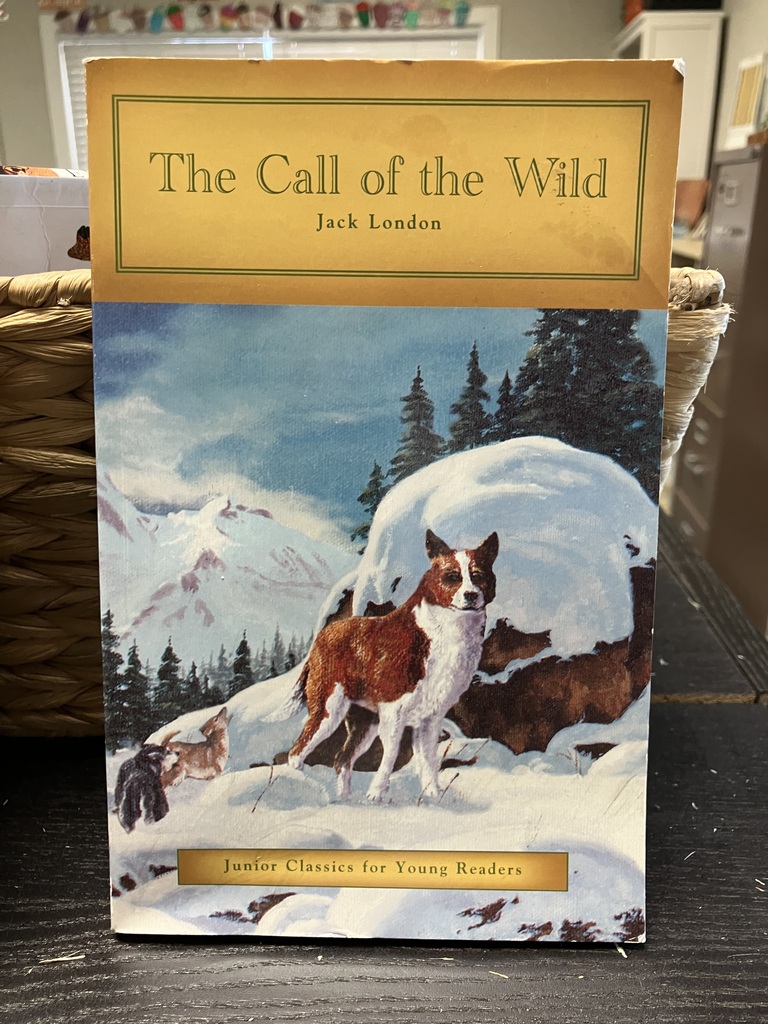 Here is a photo of the book titled "The Call of the Wild" by Jack London. On the front cover is a painting of a dog standing on the side of a snowy mountain. In the background are lots of tree and an even bigger mountain covered in snow. The dog is brown and white. The title and name of the author are both in a golden yellow framed box across the top of the cover.