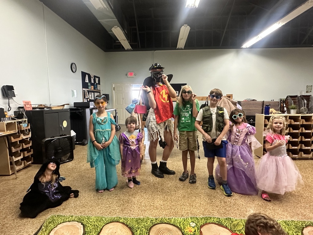 Here is a photo of a group of Afterschool students. They are all posed and looking at the camera for a photoshoot. The students are showing off their costumes they have dressed up in. Many students are wearing pink, purple, and blue princess dresses. Another student is wearing a superhero cape, there is one student wearing a safari outfit, and another wearing a witch costume. Many students are also wearing sunglasses with their costumes.