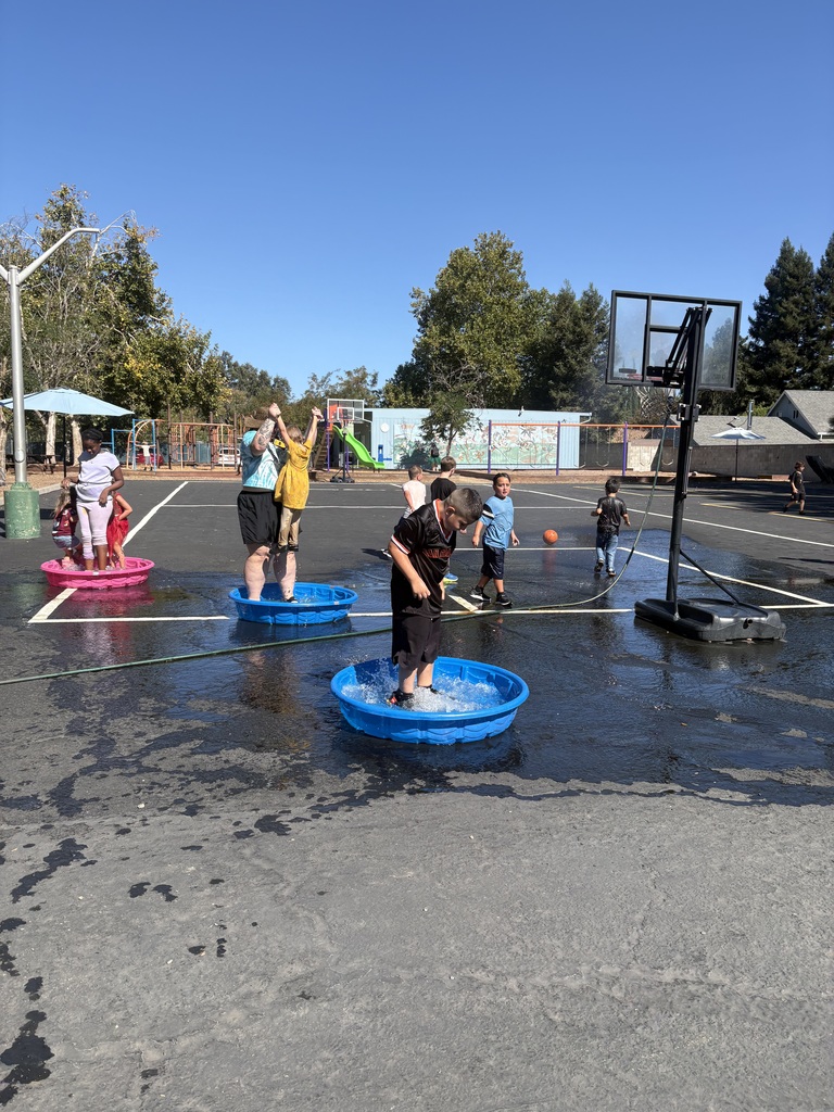 Here is a photo of a group of students playing in the water. There are students standing in small kiddie pools filled with water. The pools are set up on the blacktop. It is a bright sunny day with blue sky in the background. There are also students playing basketball on the blacktop in the background.