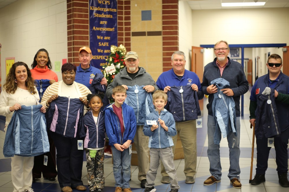 Members of the Knights of Columbus stand with school staff while presenting new winter jackets for Wilkinson County students. Several jackets are displayed, highlighting the donation for families across the district.