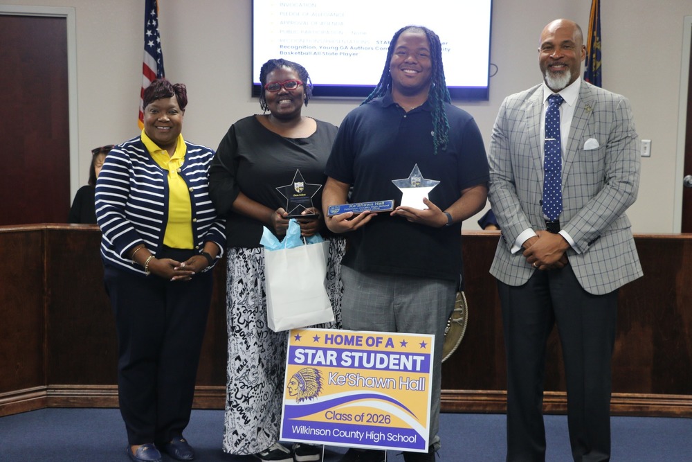 Four individuals stand together in a boardroom during a recognition ceremony. At the center, a student holds a star-shaped award and a plaque, standing behind a sign that reads “Home of a STAR Student – Ke’Shawn Hall, Class of 2026, Wilkinson County High School.” To the student’s left, a woman holds a matching award and a gift bag. Two additional adults stand on either side, all smiling toward the camera. An American flag and a presentation screen are visible in the background.