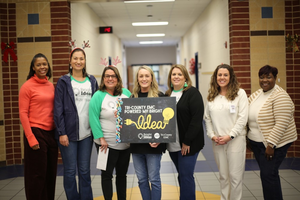 A smiling Wilkinson County Primary School teacher, Mrs. Ashley Starley, stands holding a Tri-County EMC Bright Idea Grant certificate. She is photographed inside the school with colorful student artwork in the background.