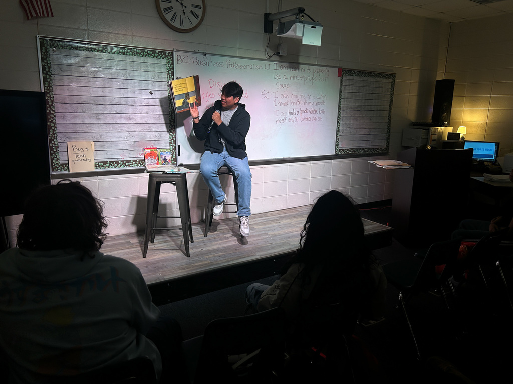 High school students in Business & Technology class practice public speaking by using props and a microphone while reading childhood books to engage their classmates.