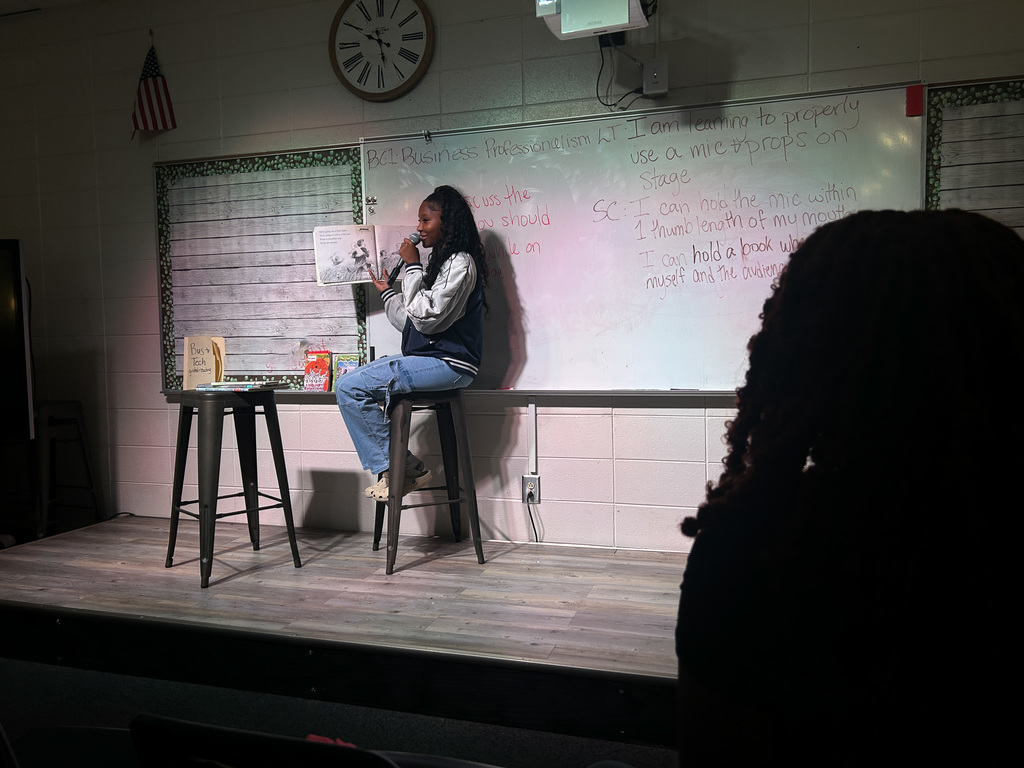 High school students in Business & Technology class practice public speaking by using props and a microphone while reading childhood books to engage their classmates.