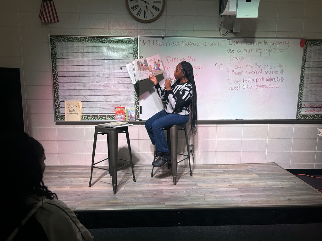 High school students in Business & Technology class practice public speaking by using props and a microphone while reading childhood books to engage their classmates.