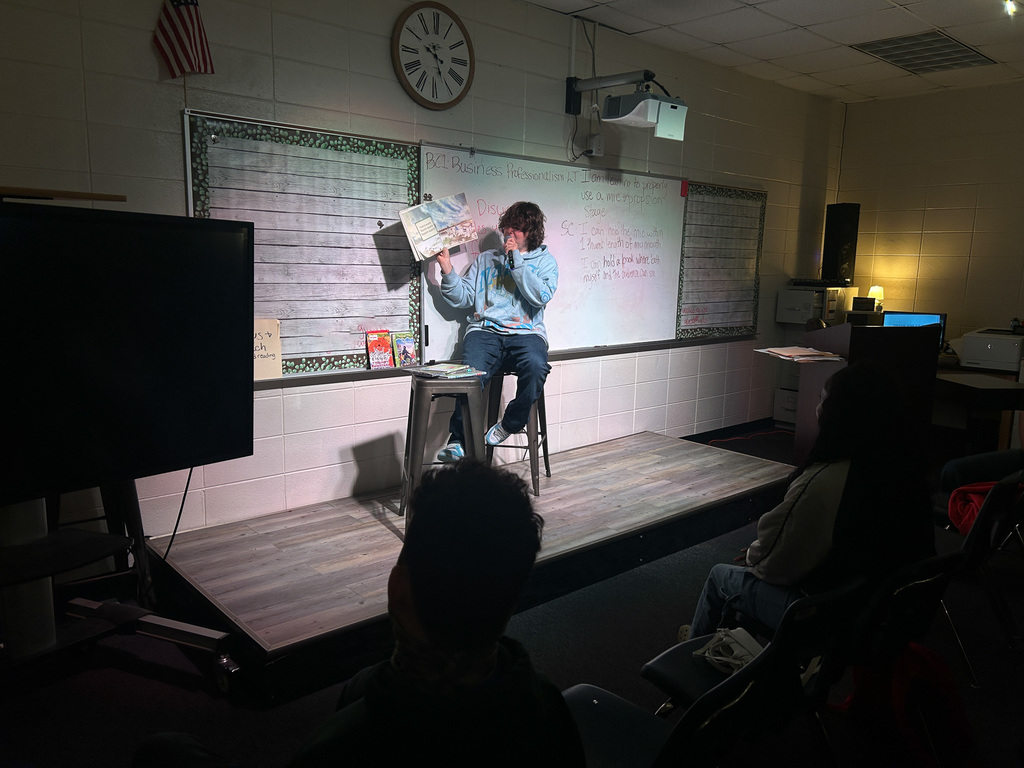High school students in Business & Technology class practice public speaking by using props and a microphone while reading childhood books to engage their classmates.
