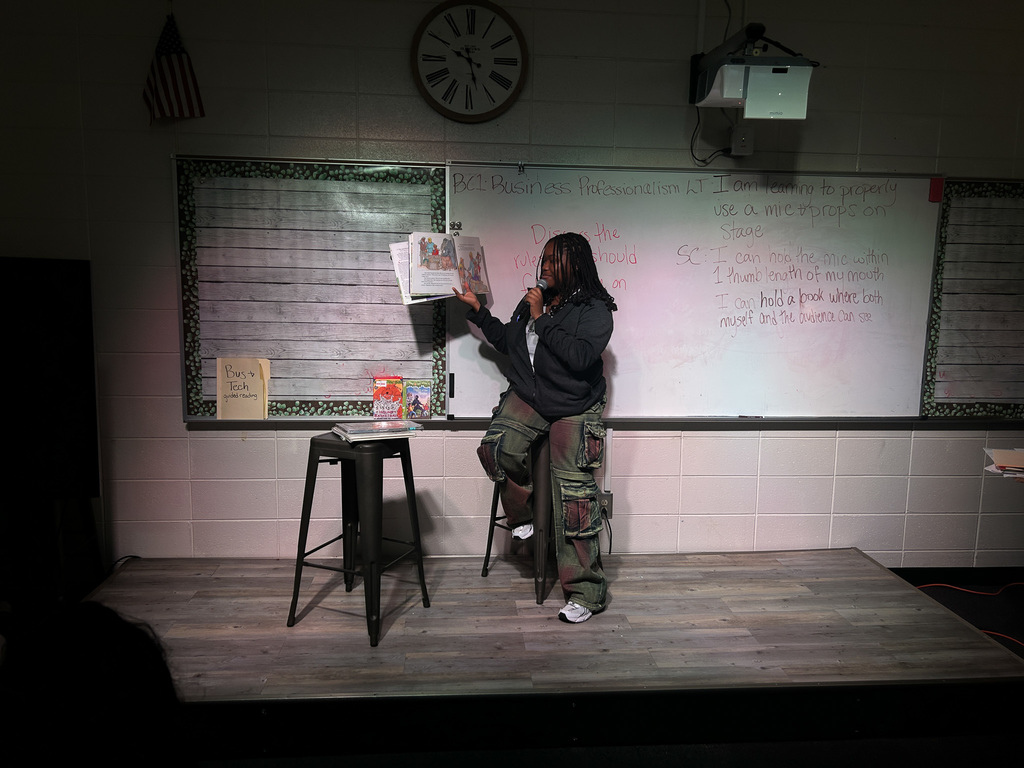 High school students in Business & Technology class practice public speaking by using props and a microphone while reading childhood books to engage their classmates.