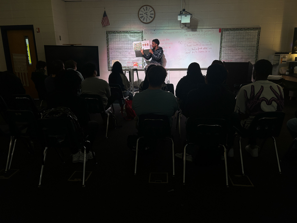 High school students in Business & Technology class practice public speaking by using props and a microphone while reading childhood books to engage their classmates.