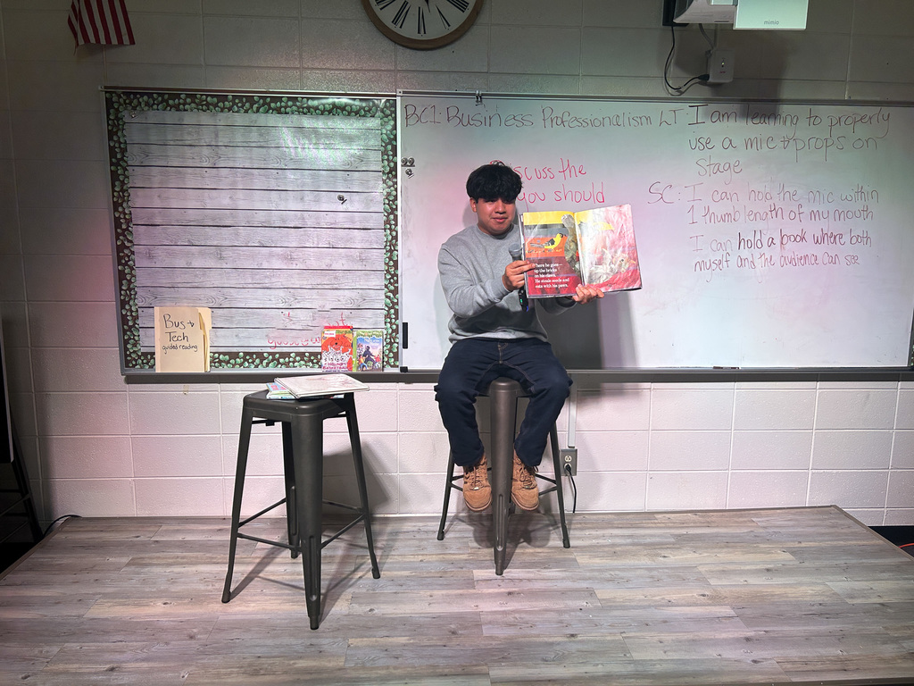 High school students in Business & Technology class practice public speaking by using props and a microphone while reading childhood books to engage their classmates.