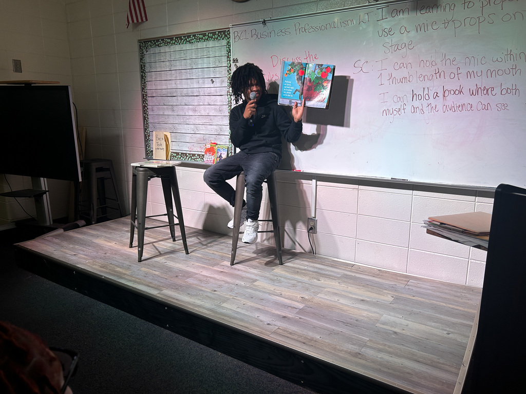 High school students in Business & Technology class practice public speaking by using props and a microphone while reading childhood books to engage their classmates.