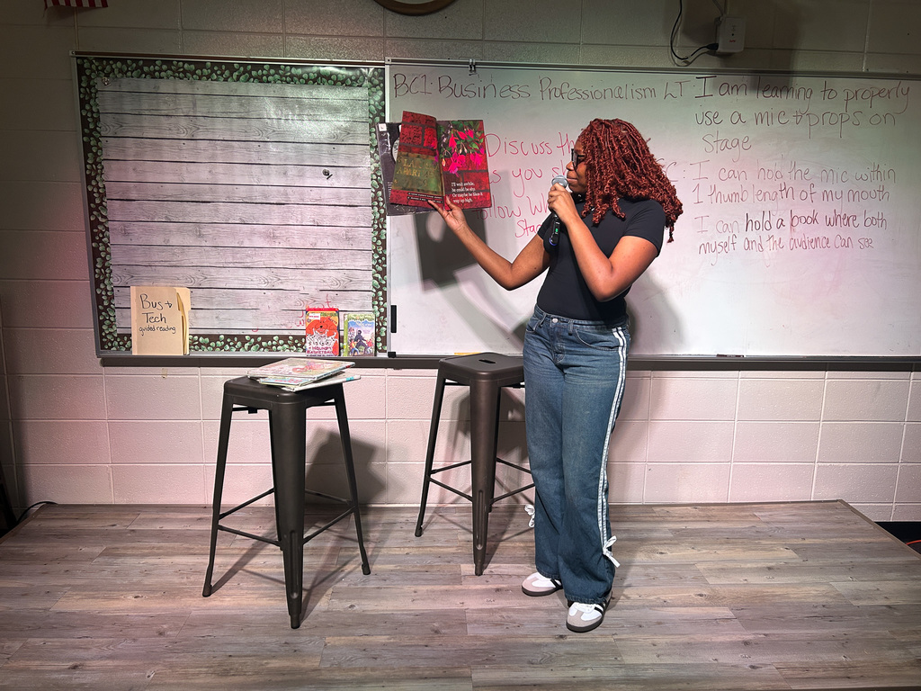 High school students in Business & Technology class practice public speaking by using props and a microphone while reading childhood books to engage their classmates.