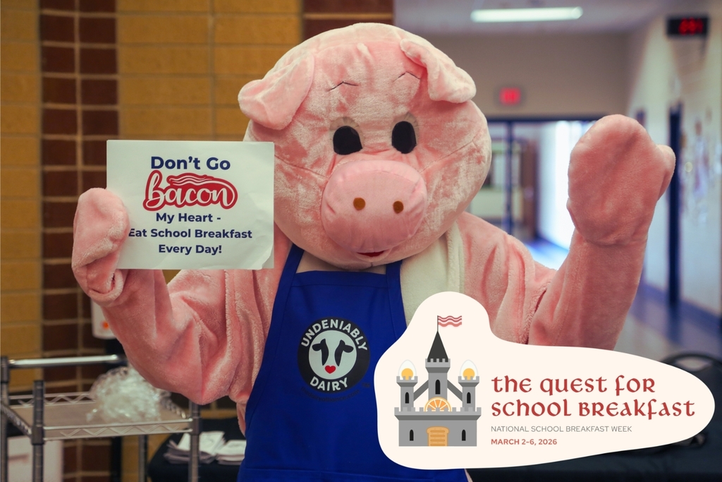 A pink pig mascot wearing a blue apron holds a sign that reads “Don’t go bacon my heart — eat school breakfast every day” inside a school cafeteria.