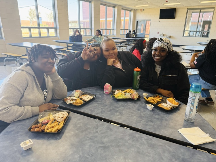 Four Wilkinson County High School students smile and pose together around a cafeteria table with their special National School Lunch Week meals, featuring funnel cake waffles, popcorn chicken, fries, and fresh produce.