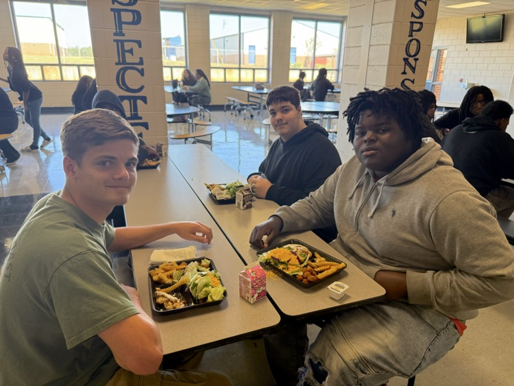 Three Wilkinson County High School students seated together in the cafeteria, smiling for a photo while enjoying their National School Lunch Week meals.