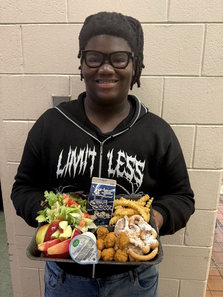 A smiling Wilkinson County High School student holds a lunch tray featuring popcorn chicken, funnel cake waffles, crinkle-cut fries, fresh apple slices, salad, ranch dressing, and milk.