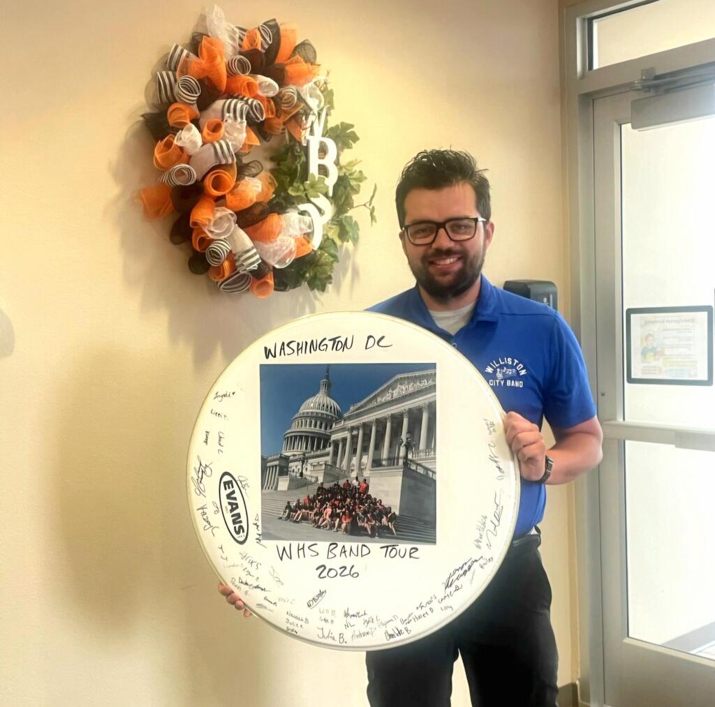 Williston High School Band Director John Bisbee holds a commemorative bass drum head created from a retired drum head and signed by band students. The display features a group photo from the 2026 WHS Band Tour to Washington, D.C., with the U.S. Capitol in the background. He stands inside the district office beneath an orange, black, and white wreath.