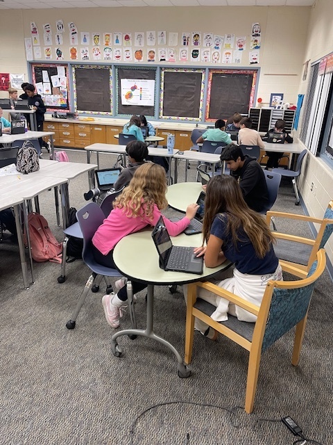 students working at a cirular table for math project while using their cromebooks to solve the case