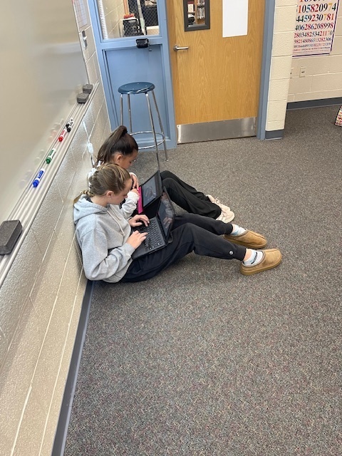 two students sitting against a wall while one is measuring on cromebook and other is typing on cromebook