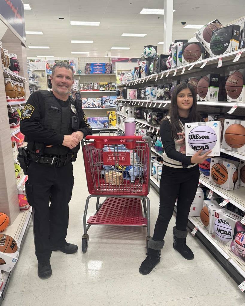 “A child stands in a sporting goods aisle at a Target store holding a boxed basketball, while a police officer stands beside a red shopping cart. Shelves filled with basketballs line both sides of the aisle.”