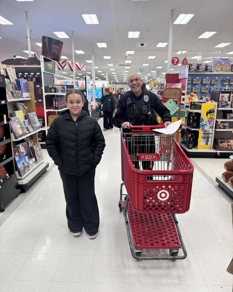 “A child stands smiling next to a police officer inside a Target store aisle. The officer, wearing a uniform and body camera, is holding a shopping cart with a clipboard, while shelves of toys and games line the aisle behind them.”
