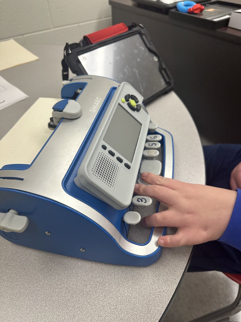A close-up of a student’s hand using a blue and silver braille embosser, pressing numbered keys while the device sits on a classroom table.