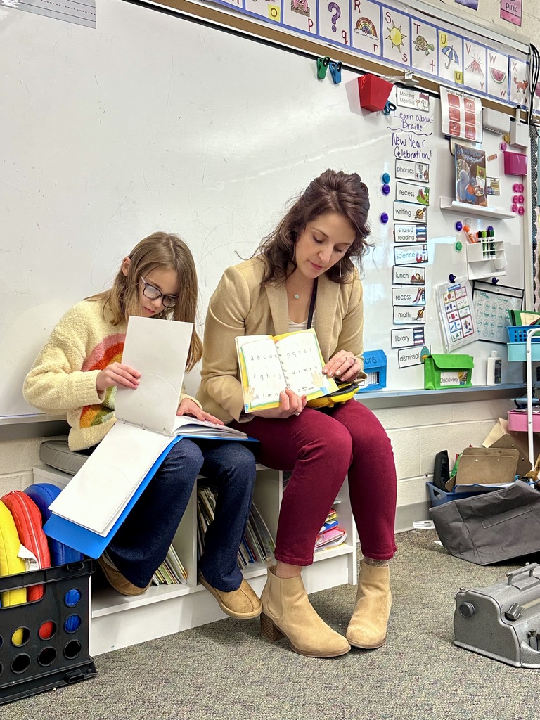 A teacher and a student sit side by side in a classroom, reading together from braille books. The teacher holds an open book while the student turns a page, with classroom materials and shelves in the background.