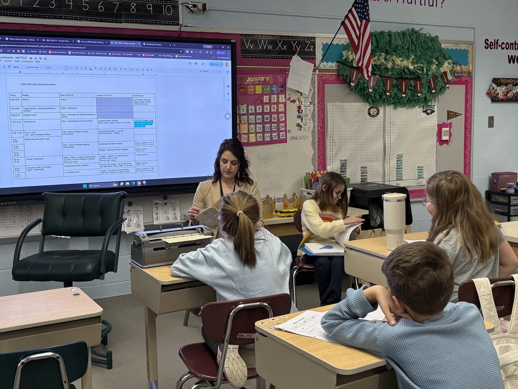 A teacher leads a small group lesson in an elementary classroom using braille materials, while students sit at desks and follow along. A smartboard and classroom decorations are visible behind them.