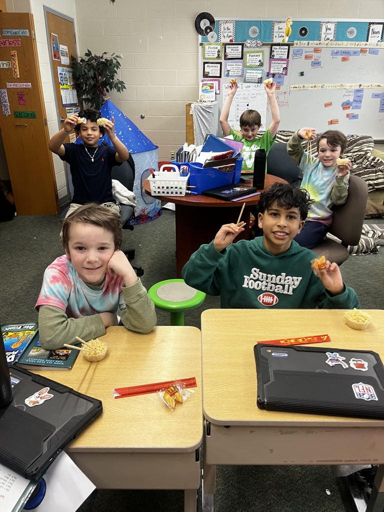 Two students sitting at desk while holding fortune cookies and chopsticks infront of small bowl of noodles  while 3 other kids are in the back doing the same thing at a table 