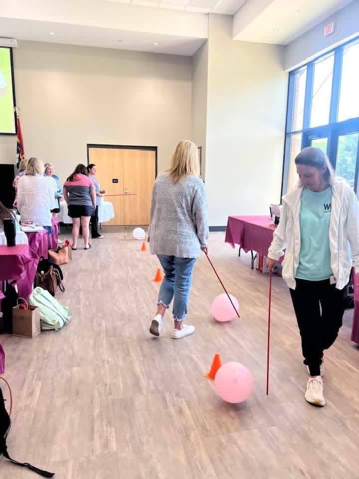 Two women with canes and balloons navigate a room with tables and chairs, participating in a guided obstacle course.