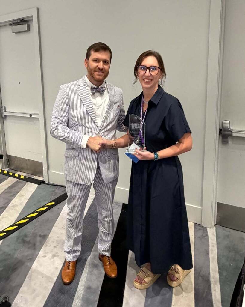 Man and woman standing next to each other, holding an award, with a white wall and striped floor behind them.