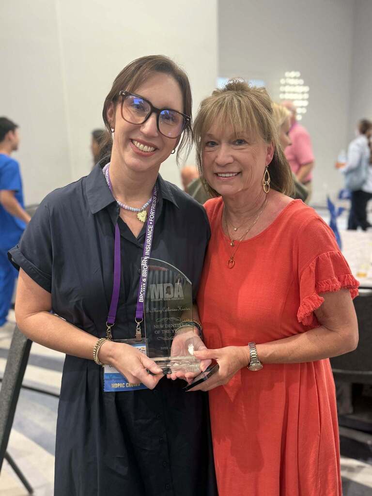 Two women standing, one holding an award, both smiling. Background shows a conference room with people.