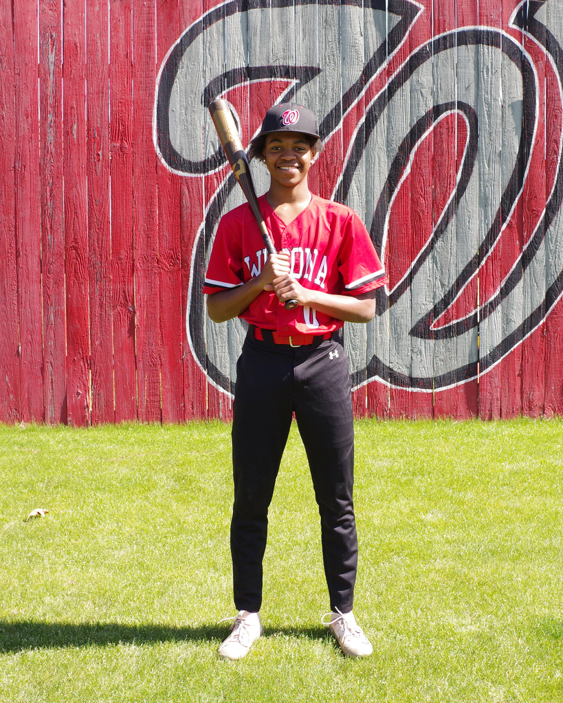 Person in red uniform, holding a baseball bat, standing on a green field in front of a red wall.