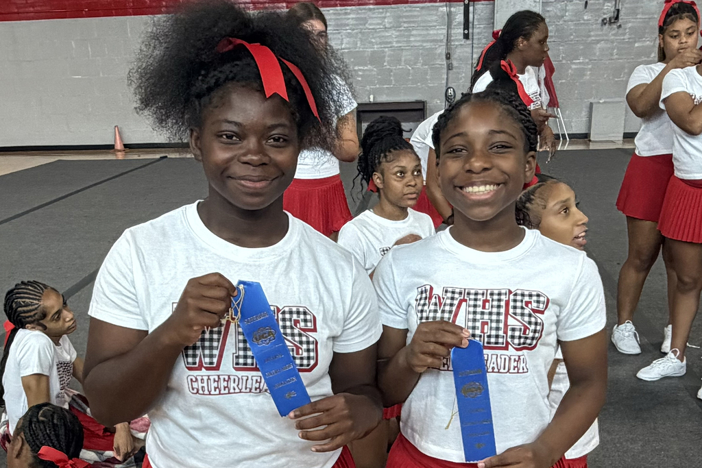 Two girls hold blue ribbons in front of a cheering squad. One girl wears a red bow.