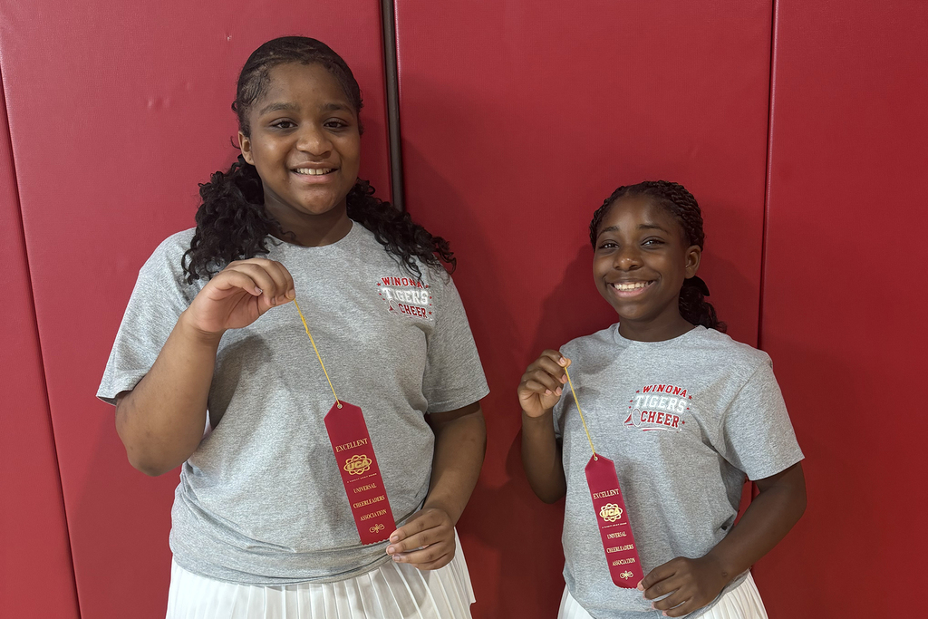 Two young girls stand next to each other, both wearing gray shirts. They hold red ribbons and smile for a photo.