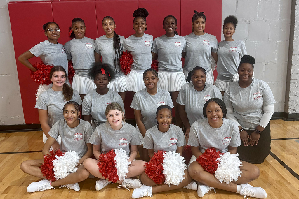 A group of young women in matching uniforms stand and sit on a basketball court with red wall behind.