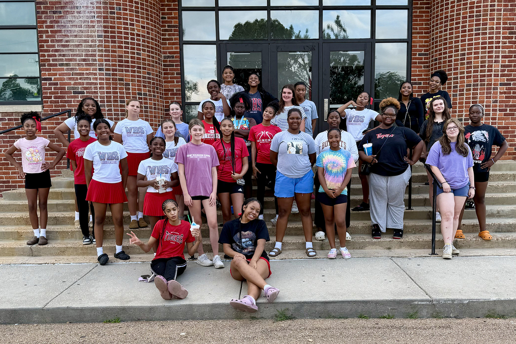 A group of people in various colored shirts and shorts pose on the steps of a brick building.