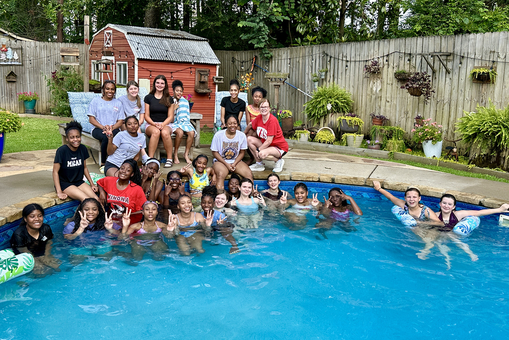 A group of people, adults and children, stand and sit in a pool and on the edge.