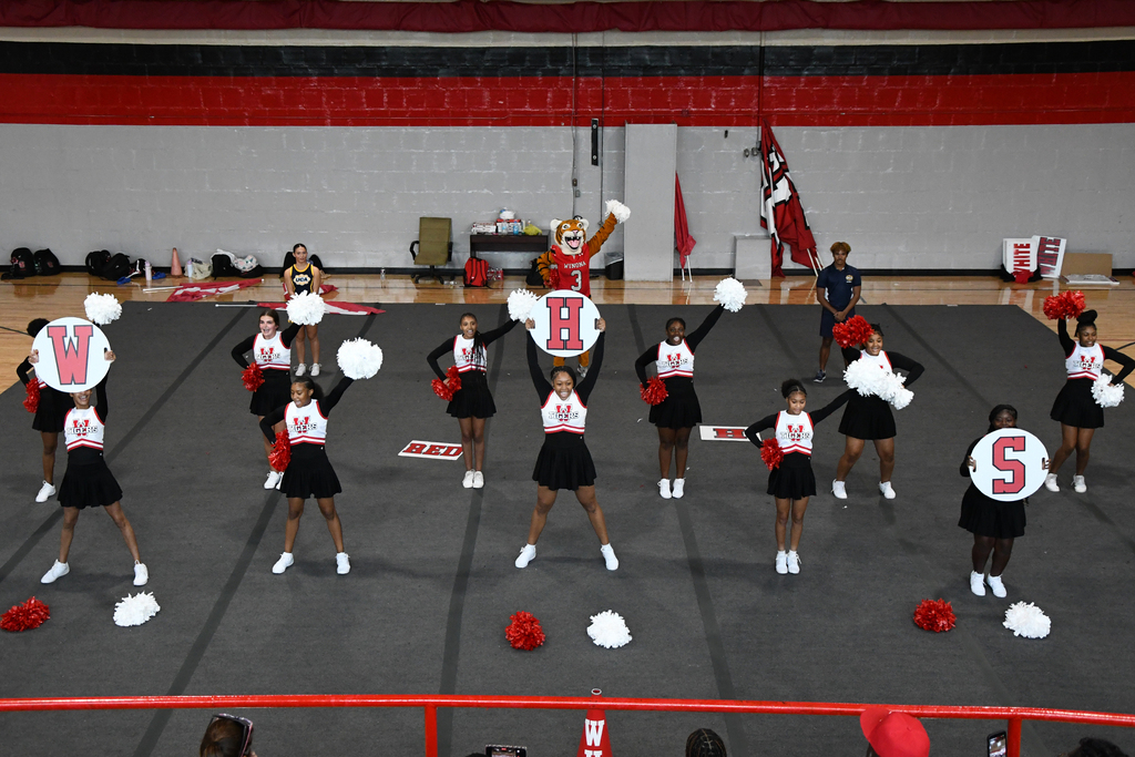 Cheerleaders in black uniforms with white shoes and pom-poms on a court. Some hold signs with letters V and S.