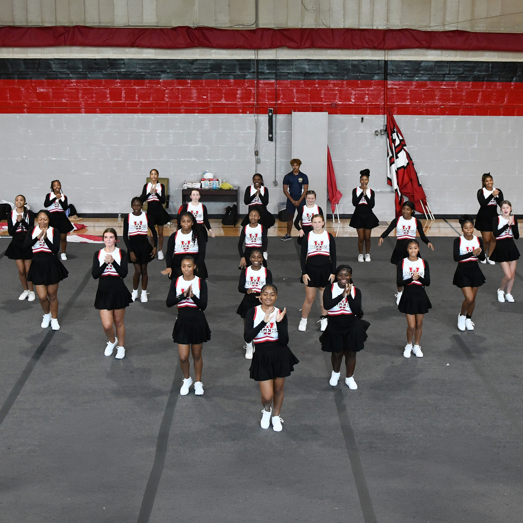 Group of cheerleaders in black uniforms performing on a gym floor. A red and black wall behind them.