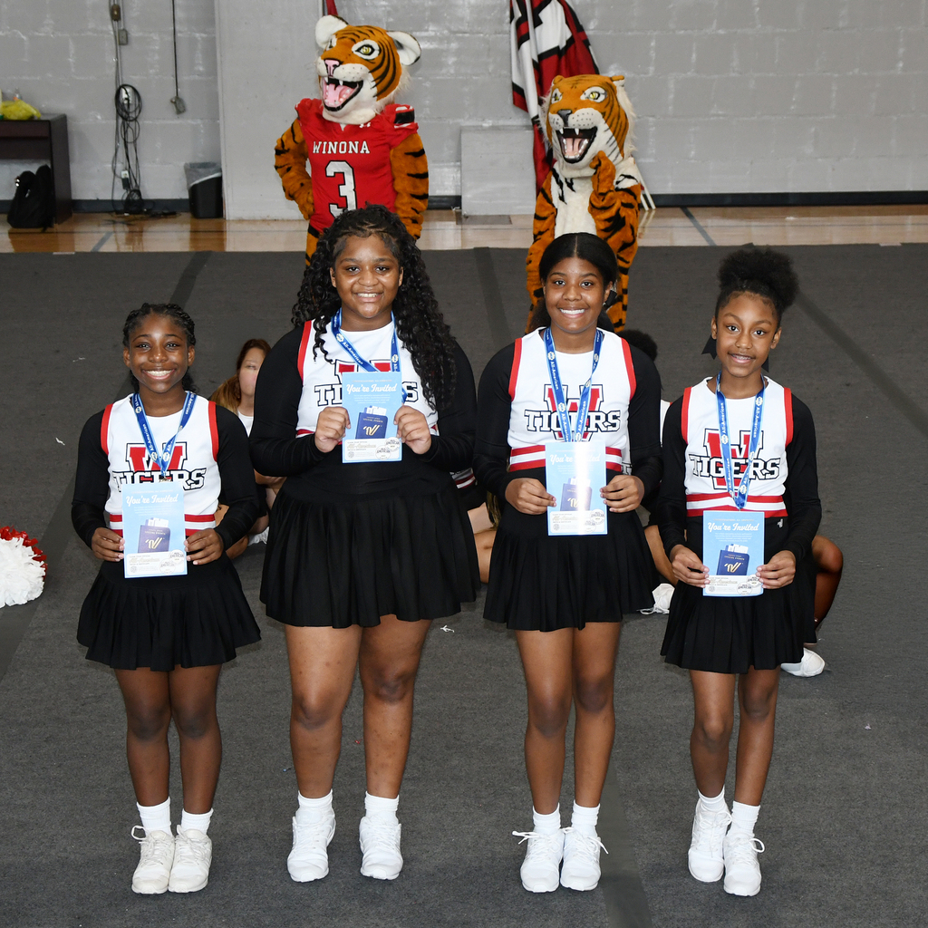 Four girls in matching uniforms with medals pose in a gymnasium with mascots in the background.