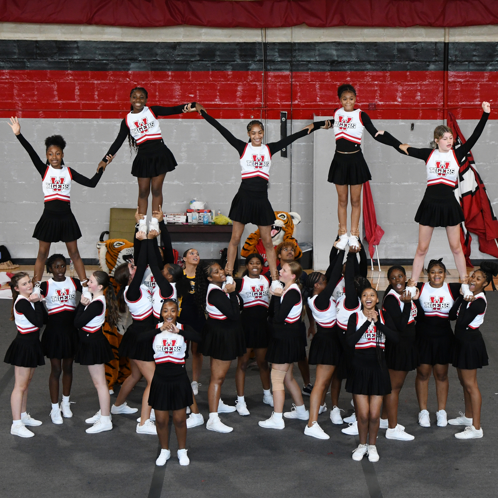 A cheerleading team forms a pyramid with three tiers, all wearing black and white uniforms. Background includes red and black wall.