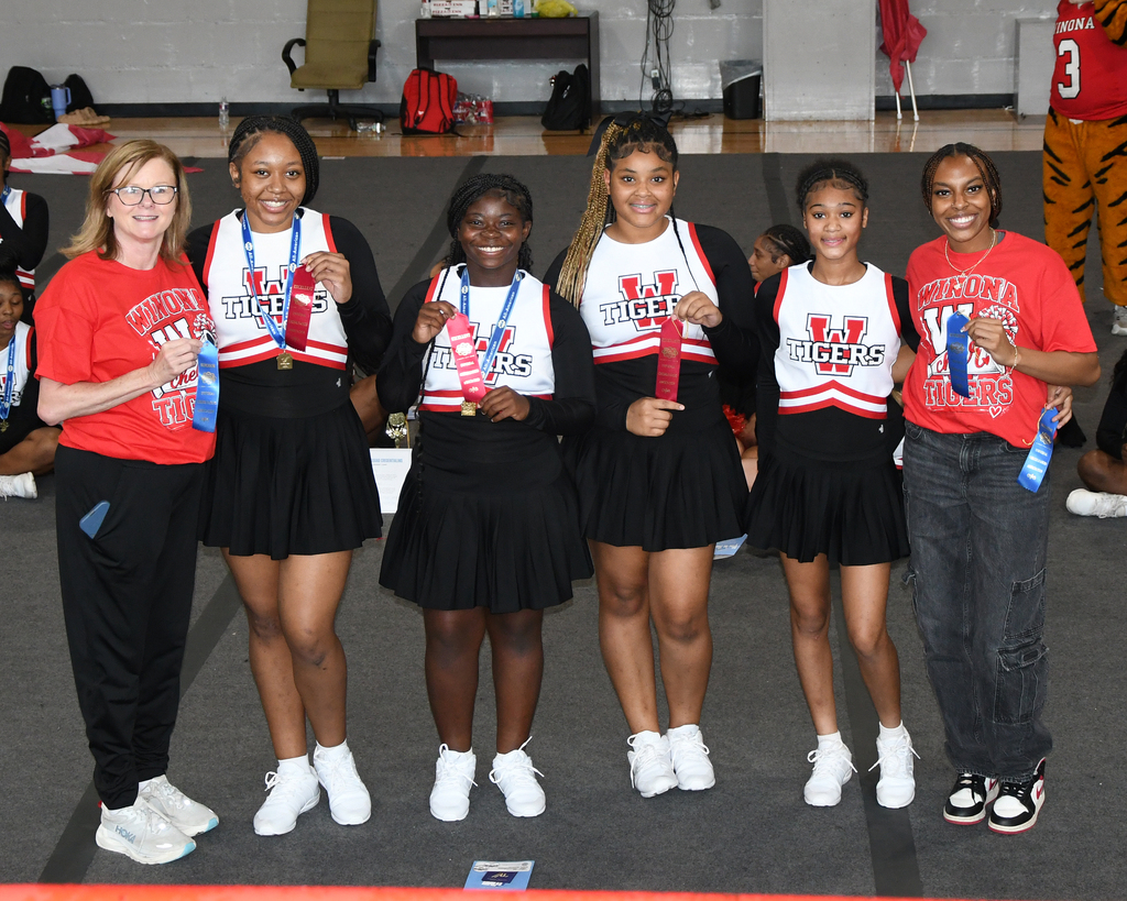 Five young women stand with their medals. They wear matching uniforms and sneakers. They are at a sports event.