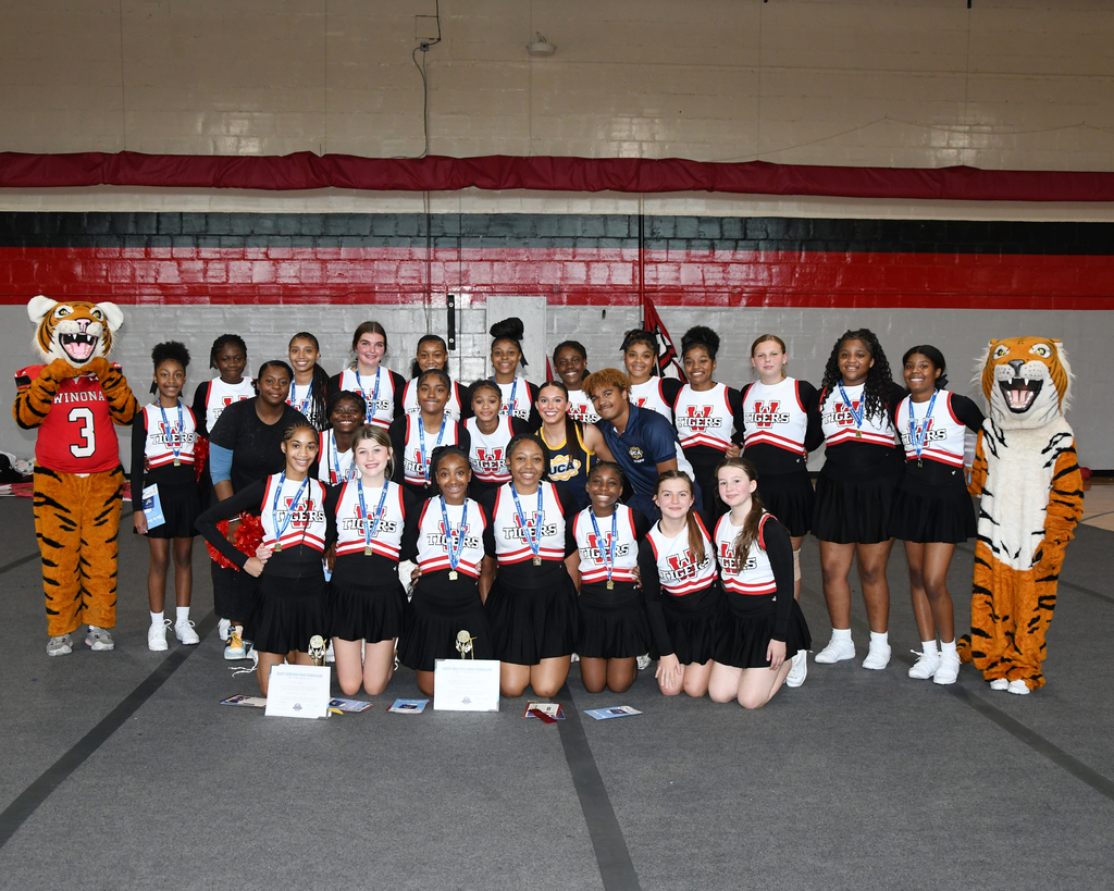 Group of cheerleaders in black and white uniforms, medals around necks, standing and kneeling on gym floor, mascots on sides.