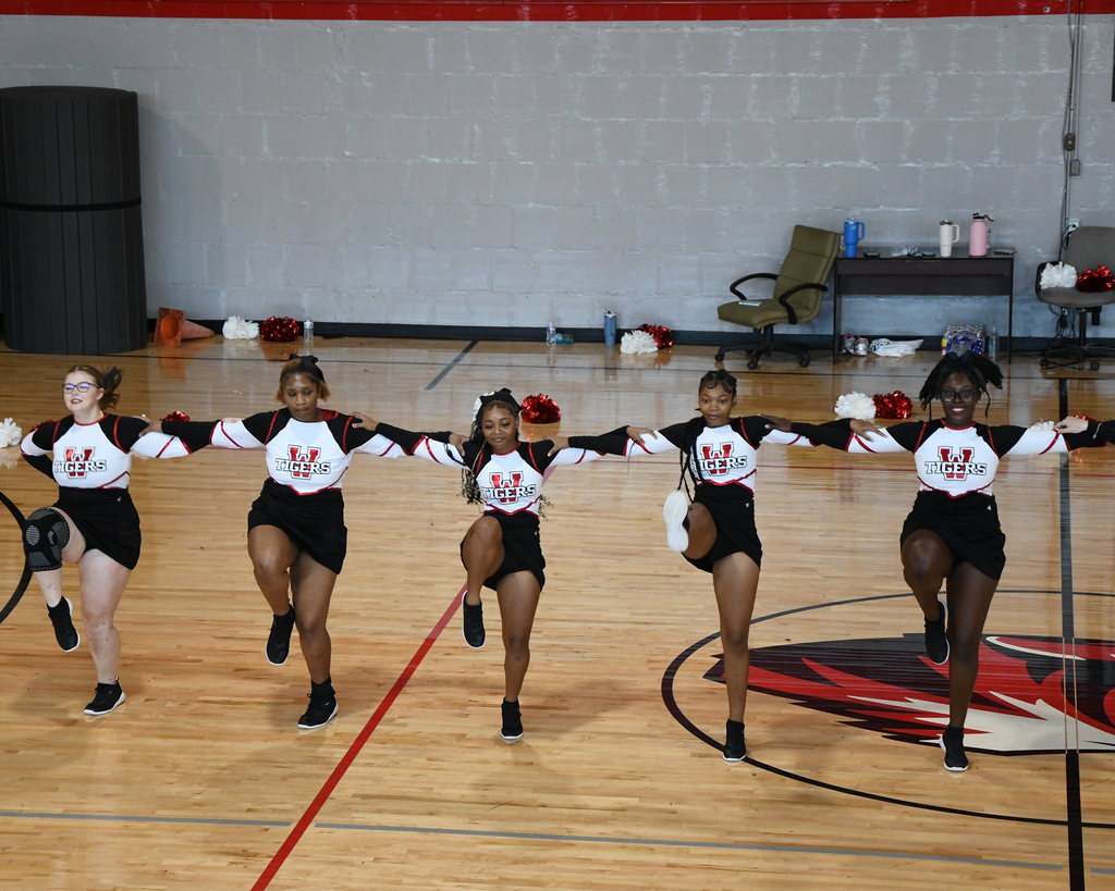 Six cheerleaders in black and white uniforms line up on a basketball court, performing a synchronized dance.