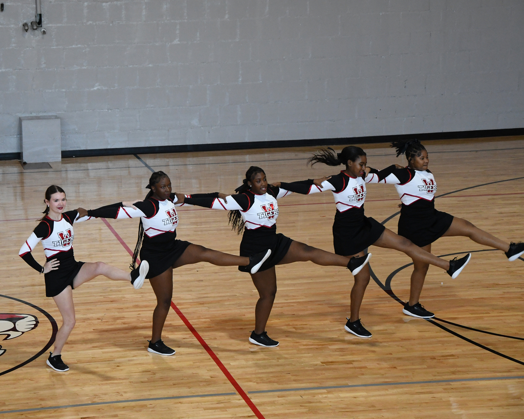 Five women in matching uniforms perform a synchronized dance routine on a basketball court.