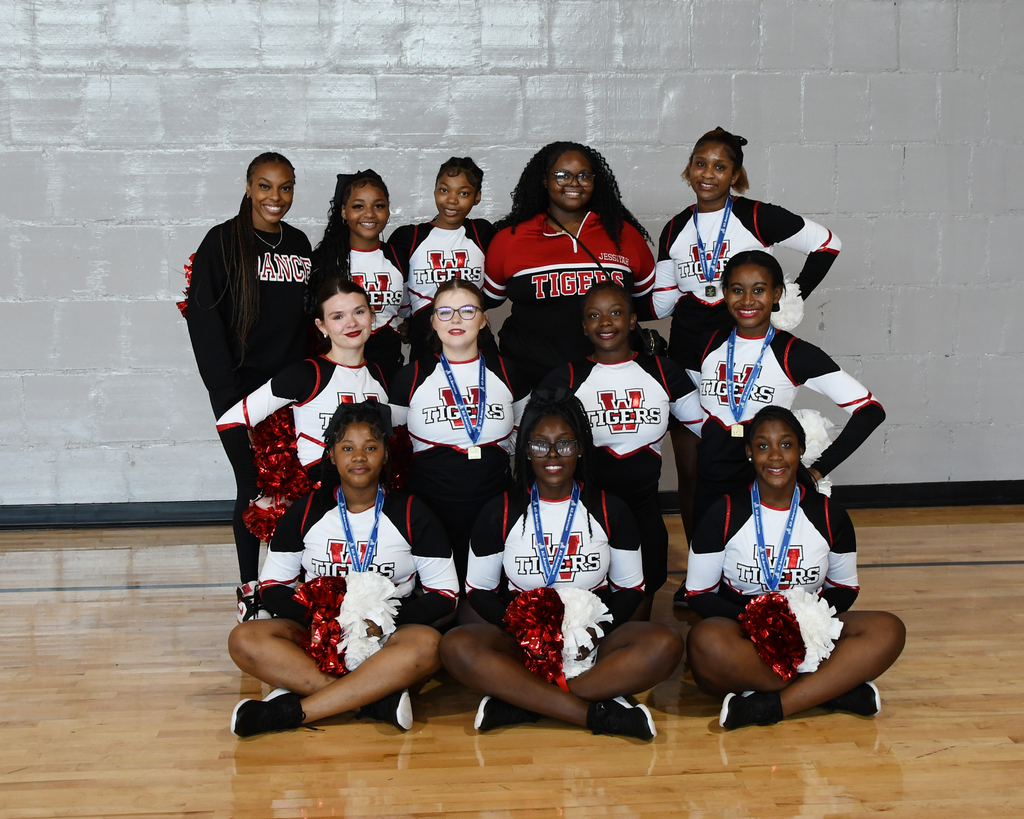Cheerleaders in uniforms, medals, and pom-poms pose for a photo in a gymnasium.