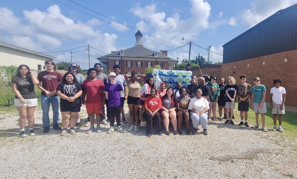 Group of people, some standing and others sitting, posing for a picture outside a brick building.