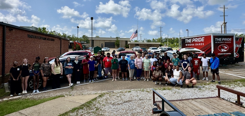 A group of people stands in a parking lot, some kneeling and smiling, with vehicles and a red truck behind.