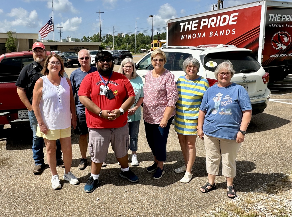 A group of people stands in a parking lot with a red truck and white SUV. Behind them is a red semi-truck with "The Pride Winona Bands" written on it.
