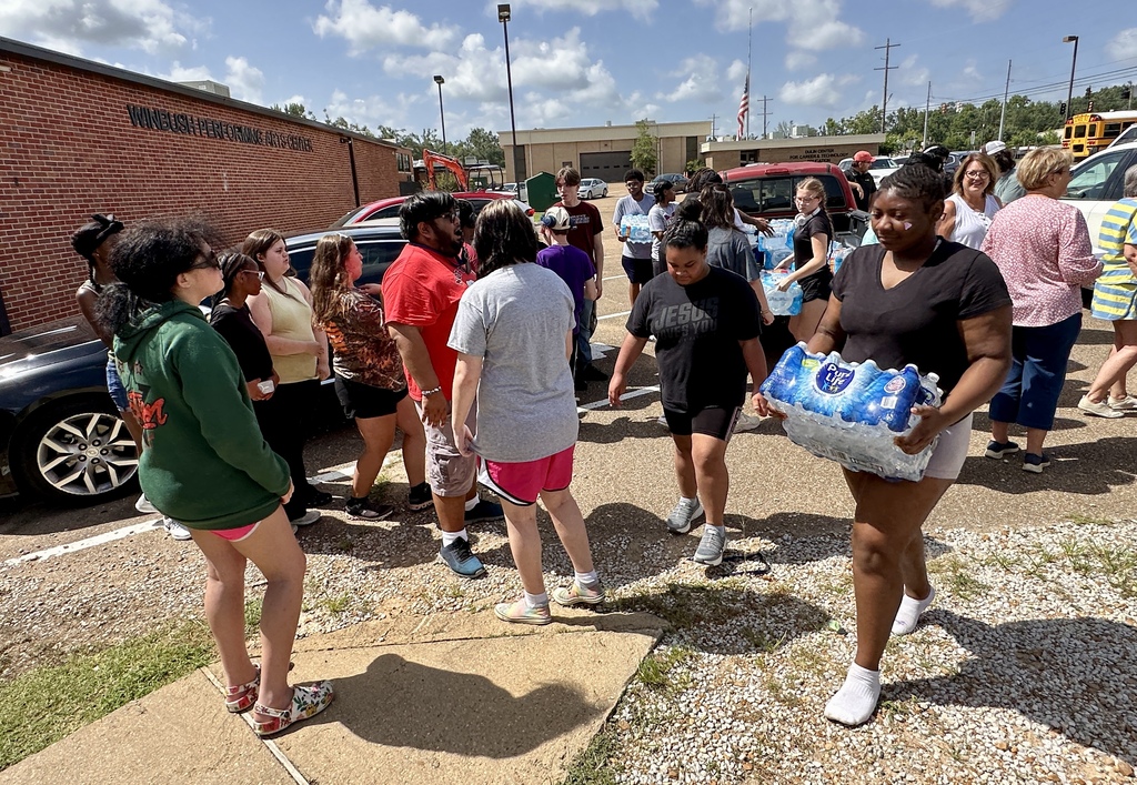 A crowd gathers outside a building; several people carry water bottles. Some wear sneakers, and others are barefoot.